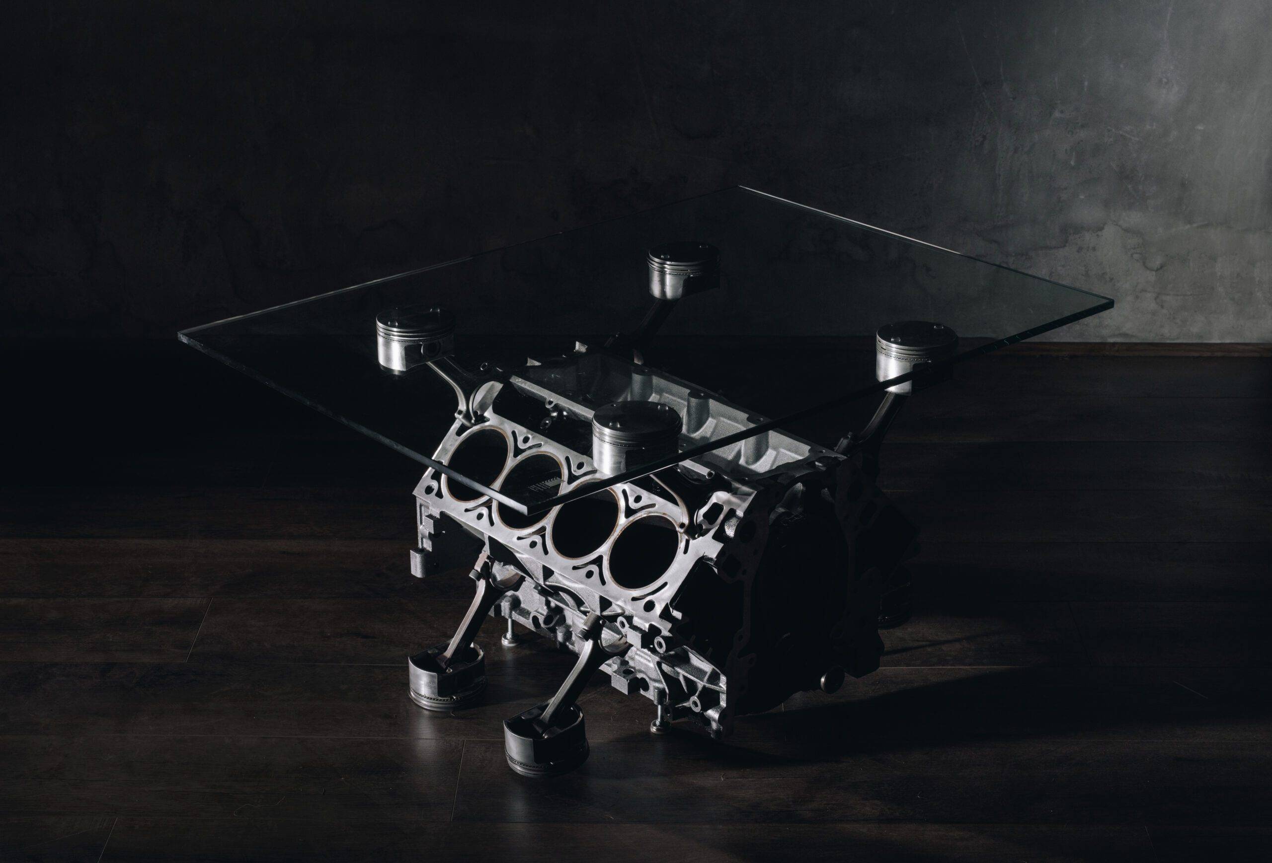 A glass coffee table with a base made from a metallic engine block, supported by eight pistons, on a dark wooden floor, Grimm Industries, Engine Coffee table. LS1 Engine coffee table.