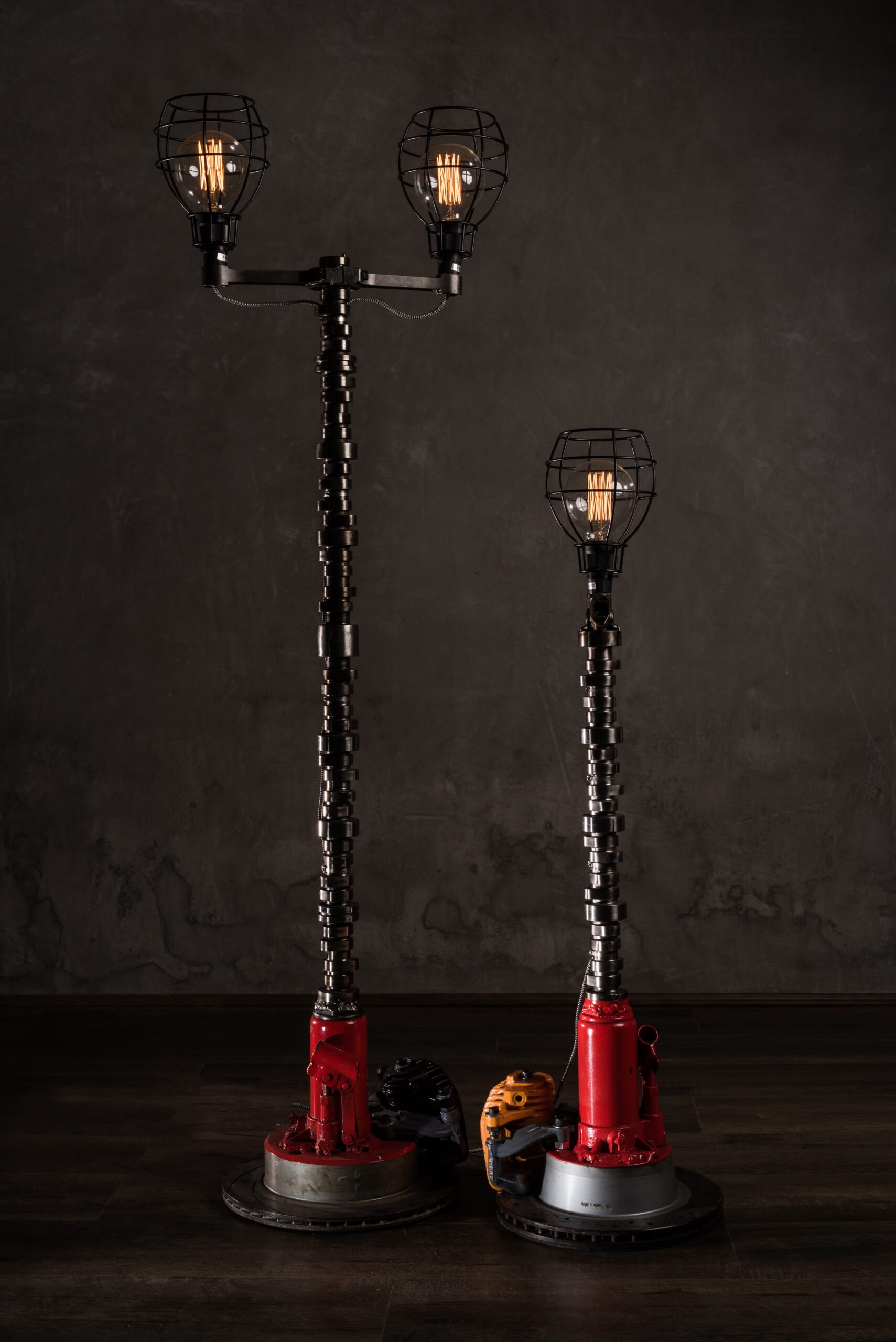 Two industrial-style floor lamps with metal cages and visible bulbs, featuring coiled metal poles and red bases on a dark wooden floor. Grimm Industries.