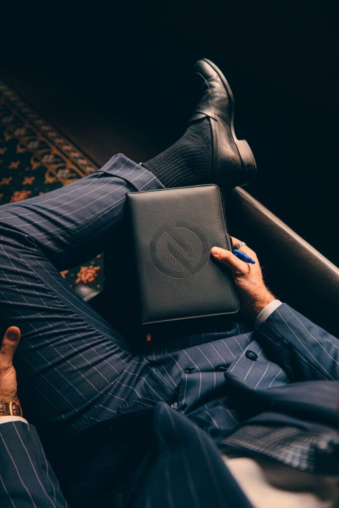 NOTA suit shop tailor, man in custom suit sitting on the chair, in suit shop setting, custom tailor, houndstooth tailors, open with phone charging and credit cards in notebook