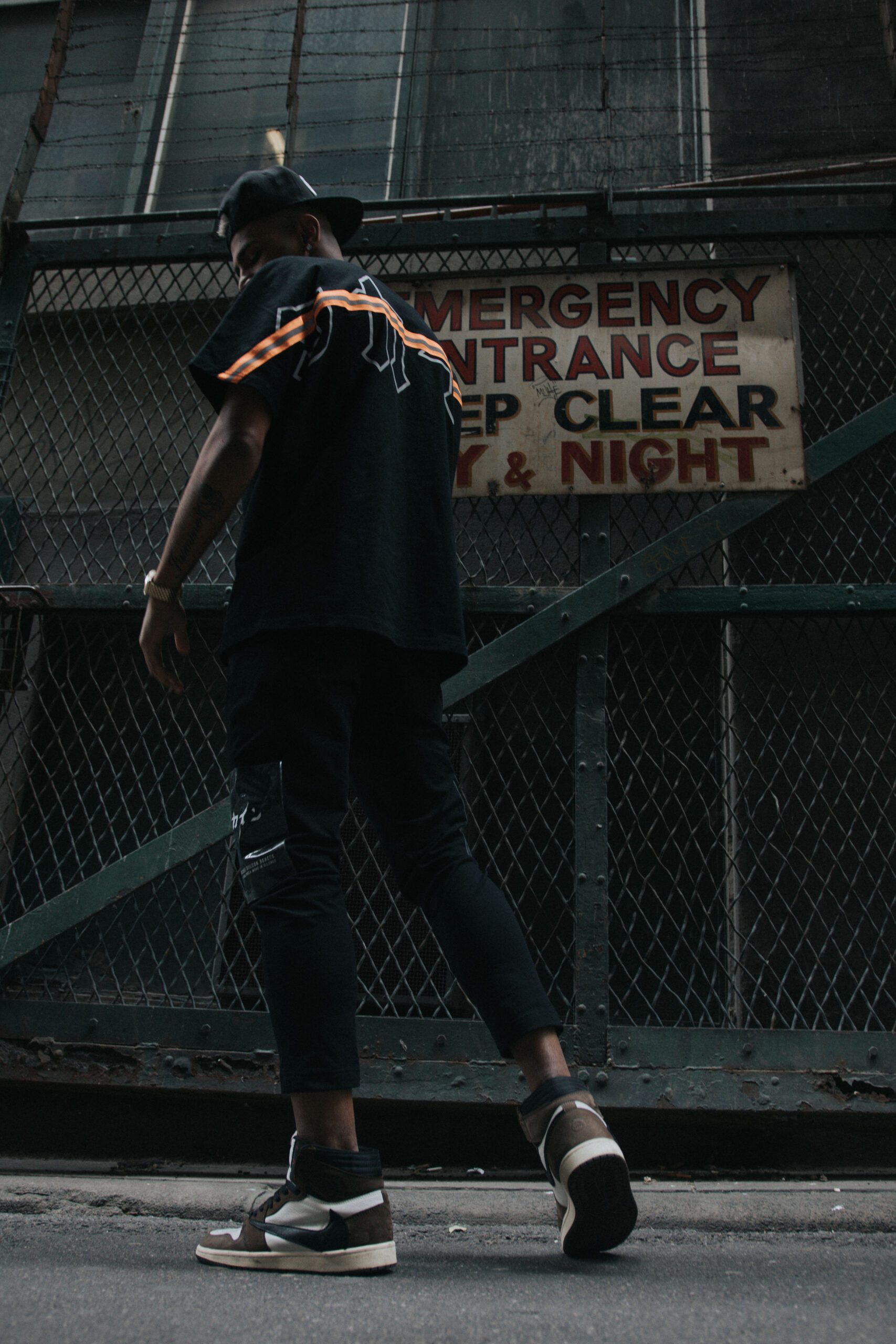 A man in a black shirt, hi vis, and travis scott sneakers walks confidently along the street, showcasing a casual urban style, wearing Kokaine